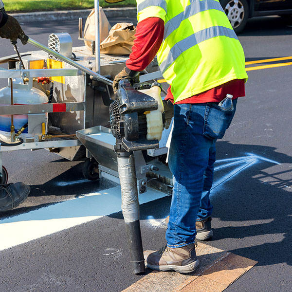 Workers are painting white road lines on an asphalt surface while traffic passes by under clear skies.