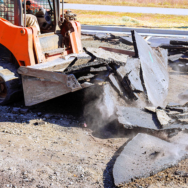 Construction skid steer machinery is breaking up asphalt in parking lot while dust rises into air