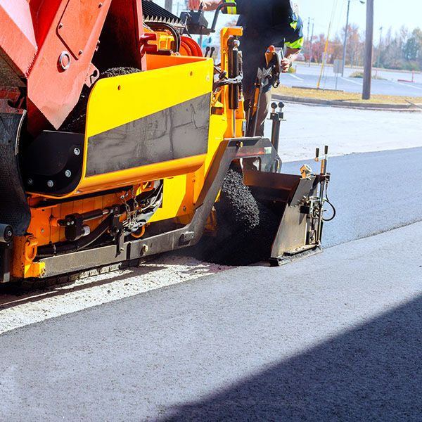Asphalt paving machine is paving asphalt on road while crew members manage process ensure safety.